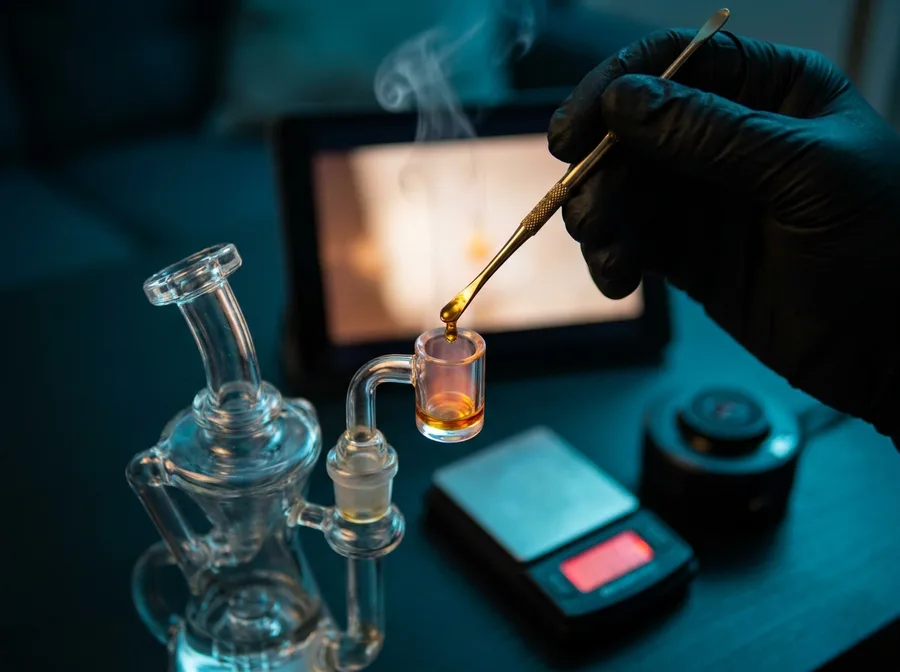 A gloved hand serving a single amber dose from a gold tool into a glowing quartz cup, the scale and camera puck watching beside it