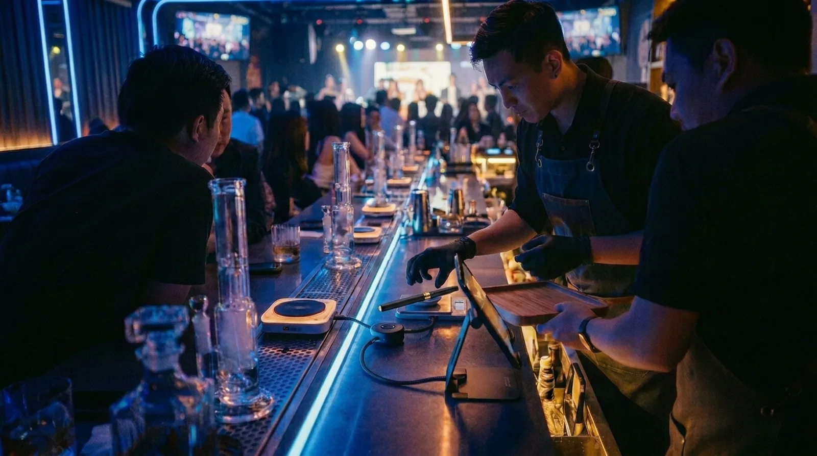 A lively lounge service bar on karaoke night — glassware down the rail, a server weighing a dose at the kit, the record keeping quietly beside the service